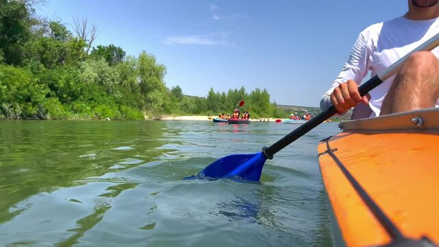 You can see how a man in a white long T-shirt and shorts is rowing with an oar with a blue blade, it then finds the camera, then moves away. A man is sitting in a kayak with an orange side, the river