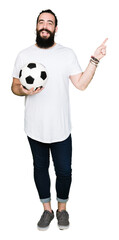 Young man with long hair and bear holding soccer football ball very happy pointing with hand and finger to the side