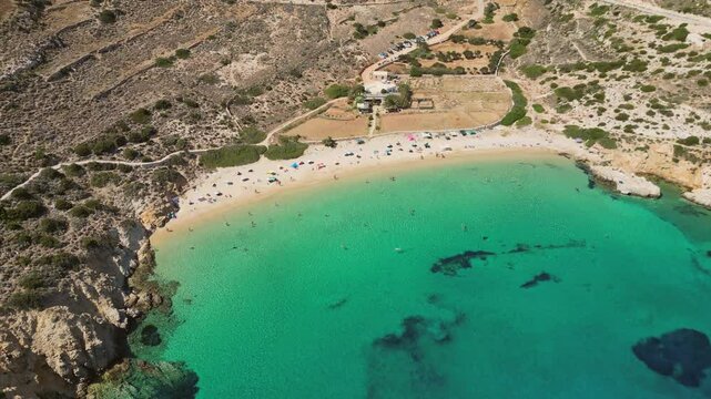 A picturesque aerial view of Kedros beach on the Greek island of Donousa. The beach is a crescent of white sand framed by turquoise waters, with a few sunbathers enjoying the idyllic setting.