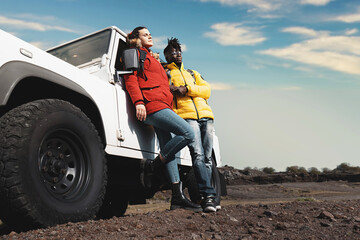 Diverse couple on adventure trip standing by off-road vehicle in rugged terrain under blue sky,...