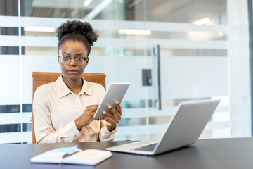 Confident professional woman using digital tablet at office desk. Modern workplace with laptop, notebook, and documents. Represents business, technology, and productivity in corporate environment.