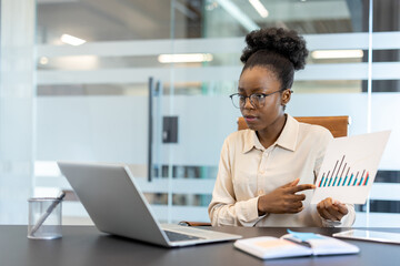 Focused businesswoman pointing at data chart in modern office setting. Confident professional using laptop for data analysis presentation. Bright office background with transparent glass elements.