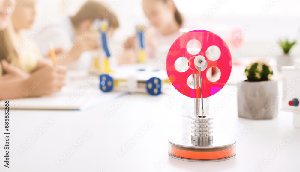 Poster A red wheel steam engine model sits on a white table in a classroom setting. The engine is made of metal and has a red plastic wheel.