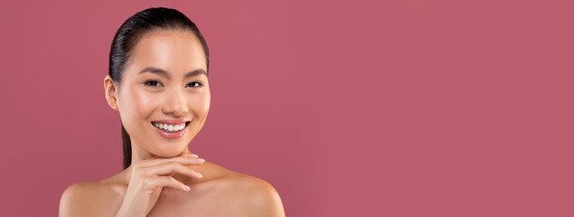 Closeup portrait of beautiful topless young chinese woman posing on purple studio background, touching her chin and smiling at camera, enjoying her smooth glow face skin, copy space