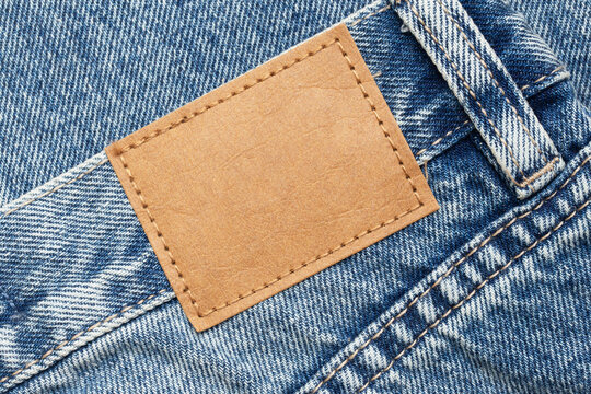 Fashion background with empty copy space for graphic design. Blank leather label tag. Blue jeans denim texture with thread sew lines. Brown clothing tag.