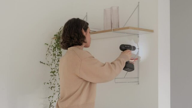 Young attractive woman hangs shelf on the wall. Female screws a screw into the shelf mount. Girl makes house cozy