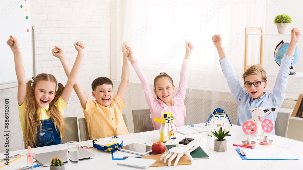 Poster four children sit at a table in a classroom, arms raised in the air, celebrating a successful roboti