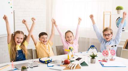 Four children sit at a table in a classroom, arms raised in the air, celebrating a successful robotics project. They are surrounded by their work, including robots, tools, and other materials