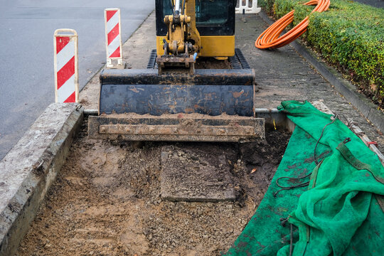 Konzept Baustelle: Frontalansicht aus der Froschperspektive auf einen Schaufelbagger an einer Stra&szlig;e mit Warnbarke, Glasfaserkabel und viel Copy Space im Vordergrund, selektiver Fokus