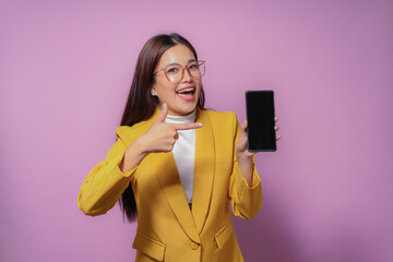 Young professional woman is holding and pointing at a smartphone with a blank screen. She is smiling and wearing a yellow blazer