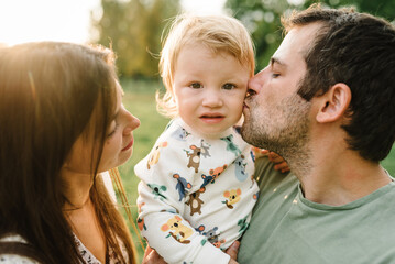 Parents kiss kid and happiness playing in green grass in park. Happy mother, father hug baby son walking in garden at sunset. Family spending time together outdoors. Children's day. Friendly family.