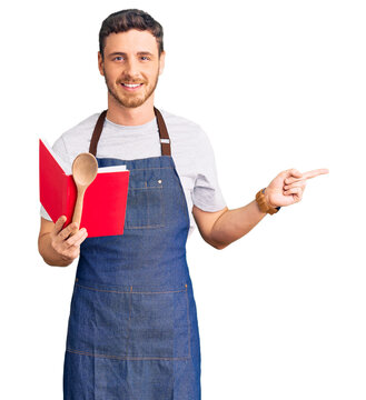Handsome young man with bear wearing professional baker apron reading cooking recipe book smiling happy pointing with hand and finger to the side