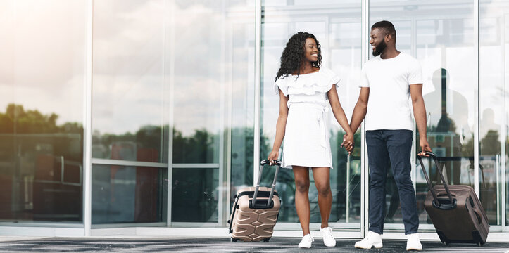 A young black couple walks hand-in-hand outside of an airport building. They are both smiling and look happy to be starting their vacation. They are each pulling a rolling suitcase behind them.