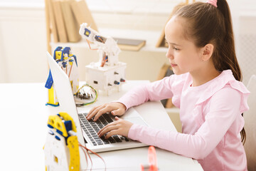A young girl wearing a pink shirt sits at a desk, concentrating as she uses a laptop. A robotic arm...
