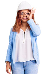 Young latin woman wearing architect hardhat doing ok gesture with hand smiling, eye looking through fingers with happy face.