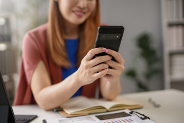 Asian businesswoman happily uses her smartphone at her office desk, connecting online with colleagues and clients. She exudes confidence and success in a modern workplace setting