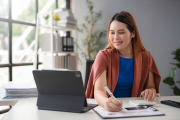 Asian accountant woman wearing a blue dress and brown blazer is smiling while working on financial documents in her bright office