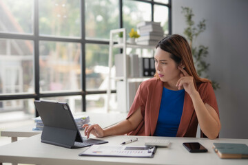 Young businesswoman is feeling tired and having a headache while working on a tablet computer in her office. She is sitting at her desk and holding her head in her hand