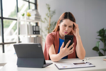 Young manager is having a headache while looking at her smartphone. She is sitting at her desk with a laptop and documents