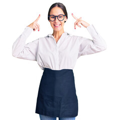 Beautiful brunette young woman wearing professional waitress apron smiling pointing to head with both hands finger, great idea or thought, good memory