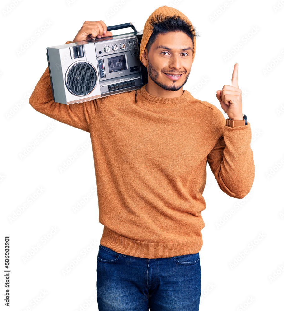 Sticker Handsome latin american young man holding boombox, listening to music pointing finger up with successful idea. exited and happy. number one.