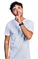 Hispanic young man with beard wearing casual white t shirt asking to be quiet with finger on lips. silence and secret concept.