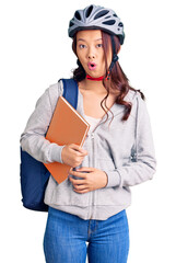 Young beautiful chinese girl wearing student backpack and bike helmet holding book scared and amazed with open mouth for surprise, disbelief face