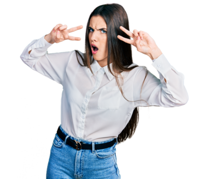 Young brunette teenager doing peace gesture close to eyes in shock face, looking skeptical and sarcastic, surprised with open mouth