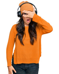 Beautiful brunette young woman listening to music using headphones peeking in shock covering face and eyes with hand, looking through fingers with embarrassed expression.