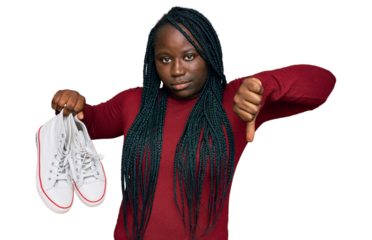 Young black woman with braids holding casual shoes with angry face, negative sign showing dislike with thumbs down, rejection concept