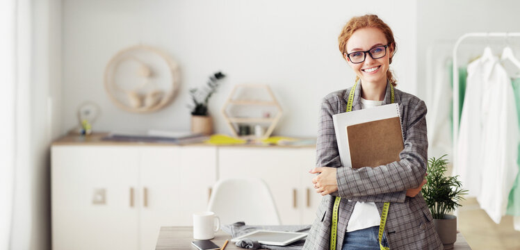 Successful Fashion Business. Smiling Young Designer In Glasses Posing In Own Dressmaking Studio Holding Sketches. Free Space - Powered by Adobe
