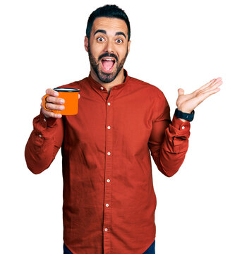 Young hispanic man with beard drinking a cup of coffee celebrating achievement with happy smile and winner expression with raised hand