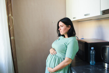 a thoughtful, sad pregnant 41-year-old woman stands in the kitchen, sideways to the camera. pregnancy after 40.