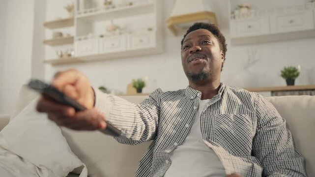 Enjoy TV show in weekend, portrait of black man with remote control in hand. Young adult African man relaxing after working days at home, watching cable or satellite television, changing channels