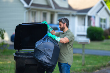 A man pushes a large plastic trash bin for weekly waste disposal schedule. Middle aged man putting...
