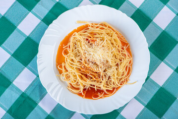 Pasta, portion of spaghetti noodles with grated cheese on a white plate on a green and white checkered tablecloth. Top view.