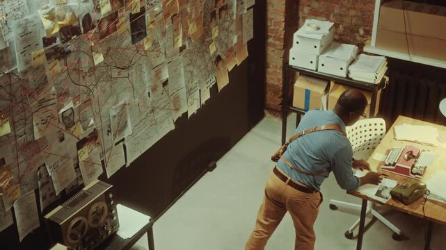 Black detective pinning paper note on investigation board covered with documents, photos and red string connections in dimly lit office at night. High angle view