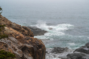 Rocky Coastline With Crashing Waves. A rocky cliff overlooks a turbulent ocean. Whitecaps crash against the jagged rocks, creating a dramatic scene. The water is a deep blue, and the sky is overcast,
