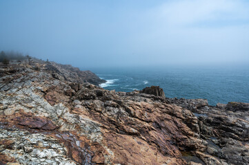 Rocky Coastline on a Foggy Day. A panoramic view of a rugged, rocky coastline overlooking the vast blue ocean. A thick layer of fog hangs in the air, obscuring the distant horizon. The rocky terrain 