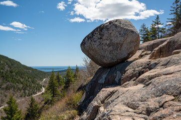 Precariously Balanced Bubble Boulder on a Mountainside Overlooking the Ocean. A large, round boulder rests precariously on a smaller rock formation, offering a breathtaking view of a winding road lea