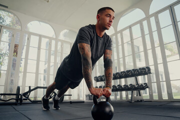 Young tattooed multiracial man in sports clothing in plank position with kettlebell at gym