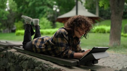 Happy girl student with curly hair in a checkered shirt uses a tablet and lies on a bench in the park