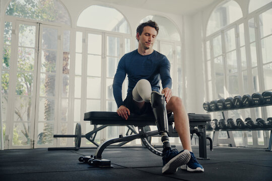 Young caucasian man in sports clothing sitting on bench press while attaching prosthetic leg at the gym