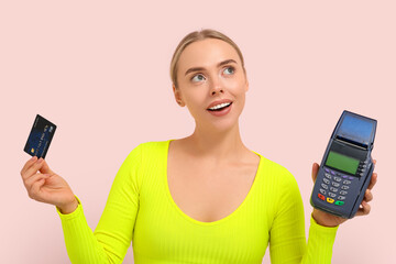 Pretty young woman with credit card and payment terminal on pink background