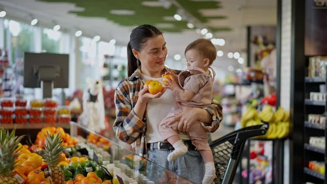 Happy brunette woman in a plaid shirt holds a small child in her arms and shows him orange fruits in a supermarket department