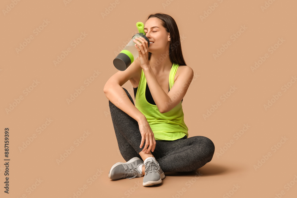 Poster young sporty woman sitting and drinking water from bottle on beige background