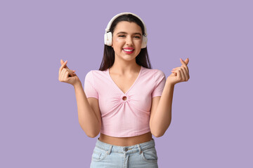 Beautiful young woman in modern headphones showing heart gesture on lilac background