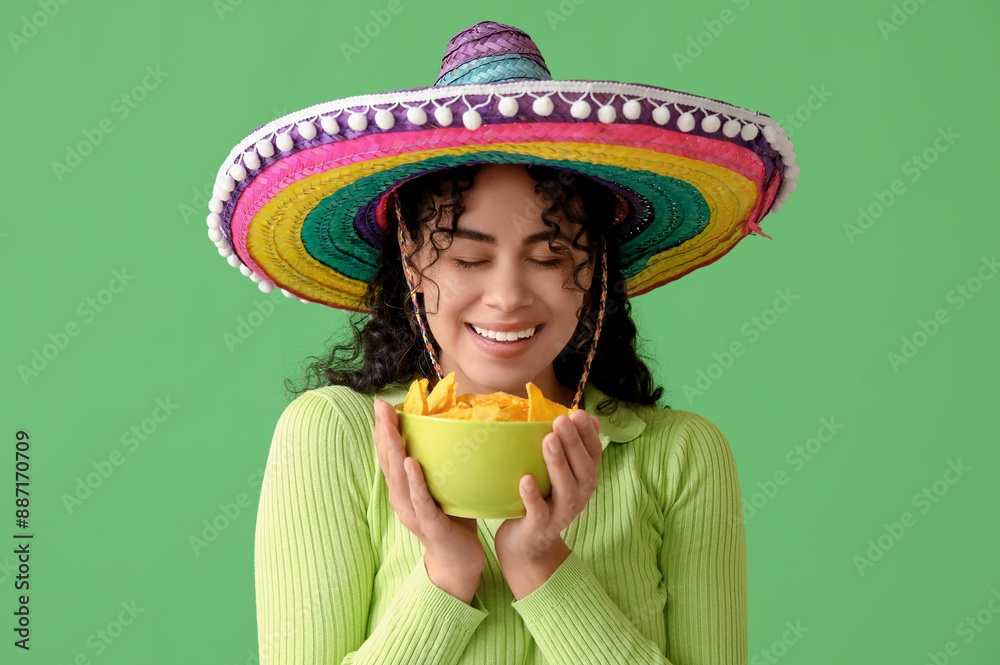 Poster Happy young African-American woman in sombrero hat holding bowl with tasty nachos on green background