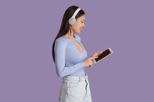Beautiful young Asian woman in headphones listening to audiobook with modern tablet computer on lilac background