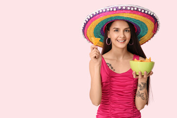 Beautiful young woman in sombrero hat with tasty nachos on pink background